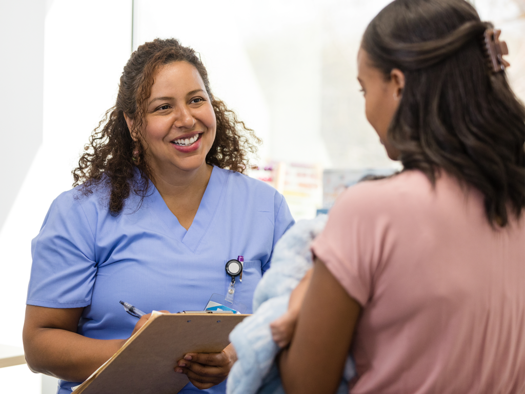 Female nurse, smiling and talking to female patient in exam room with clipboard in hand